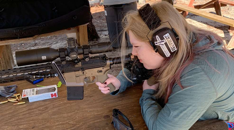Woman shooting an AR-style rifle with a Vudu scope from a bench rest at an outdoor range, with Hornady ammunition boxes visible on the table.