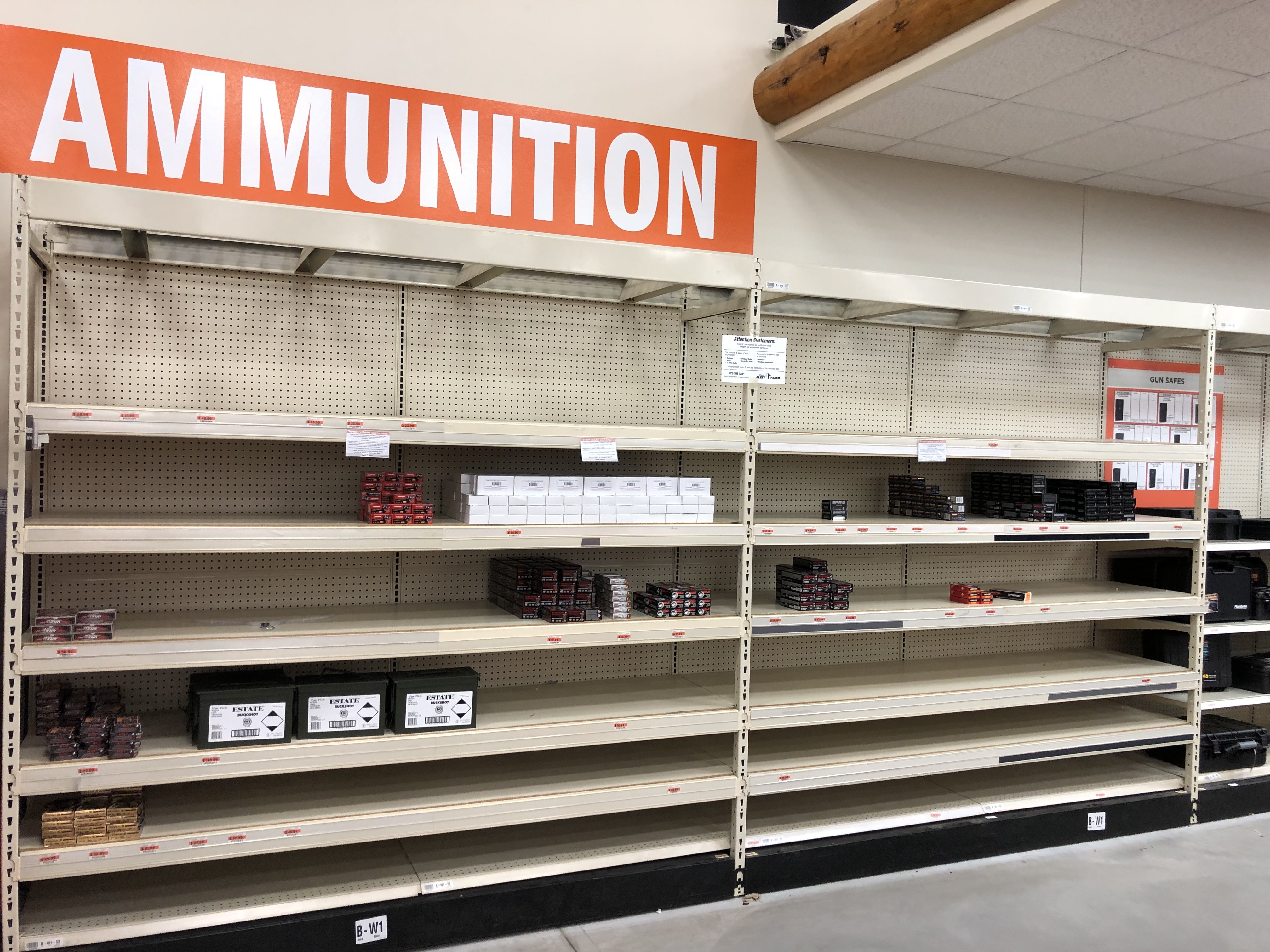 Nearly empty retail ammunition shelves with sparse boxes of handgun and shotgun ammo remaining beneath a large orange Ammunition sign.