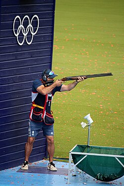 A competitive shooter in shooting vest and eye protection fires a shotgun at an Olympic clay target shooting venue with the Olympic rings visible on a wall behind him.