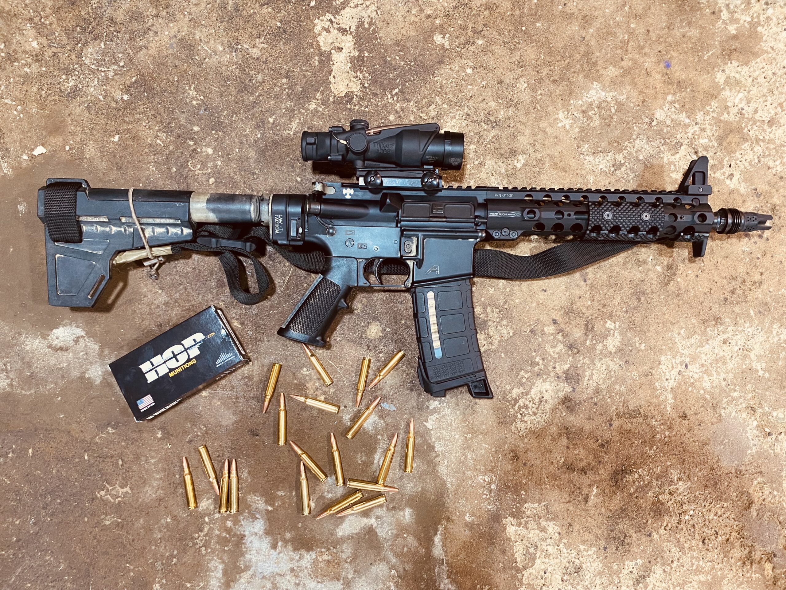 AR-pistol with Trijicon ACOG optic, pistol brace, and Magpul magazine laid on concrete next to a box of HOP Munitions and approximately 20 loose rifle cartridges.