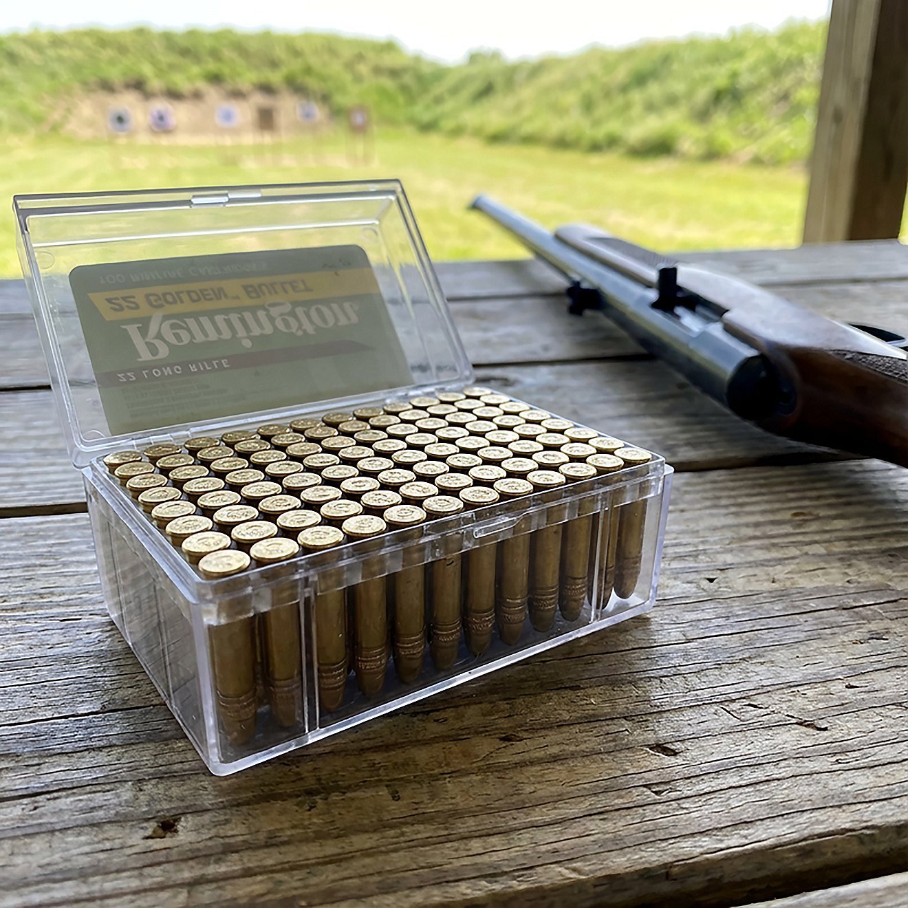 Clear plastic box holding 100 rounds of Remington .22 LR ammunition on a wooden shooting bench, with a rimfire rifle and paper targets visible in the background.