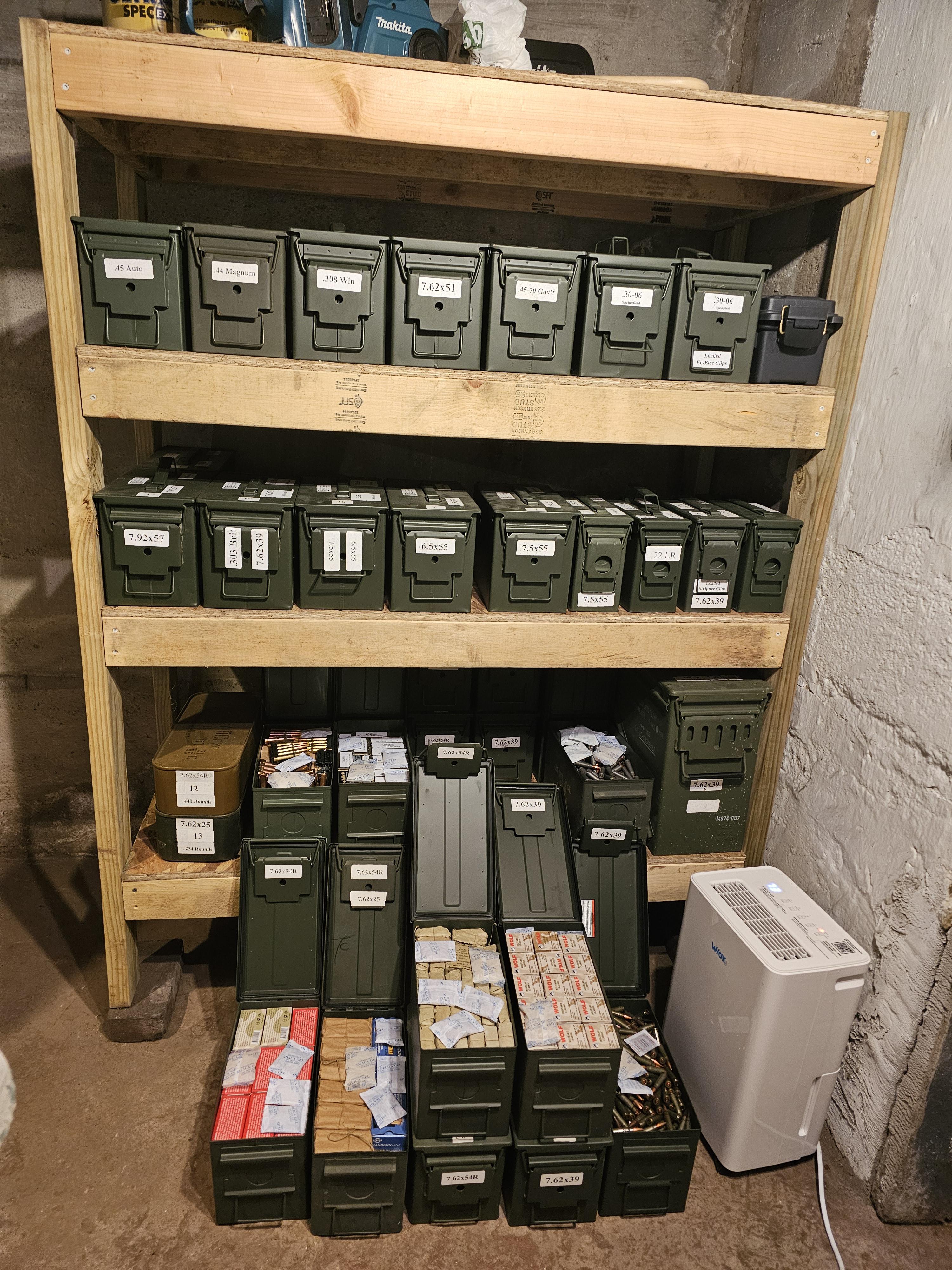 Wooden shelving unit holding dozens of labeled military surplus ammo cans organized by caliber, with loose ammunition boxes and a small dehumidifier visible on the bottom shelf.