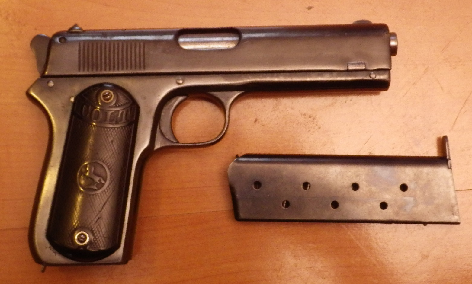Colt Model 1903 Pocket Hammerless pistol with detached single-stack magazine, displayed on a wooden surface, showing original checkered hard rubber grips with Colt medallion.